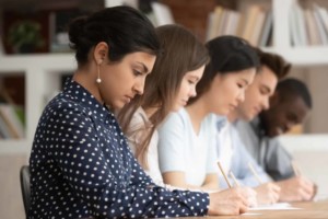 Multi-ethnic concentrated students sitting at desk holding pencils writing on papers doing task, diminishing perspective focus on indian girl study process or assessment - knowledge evaluation concept