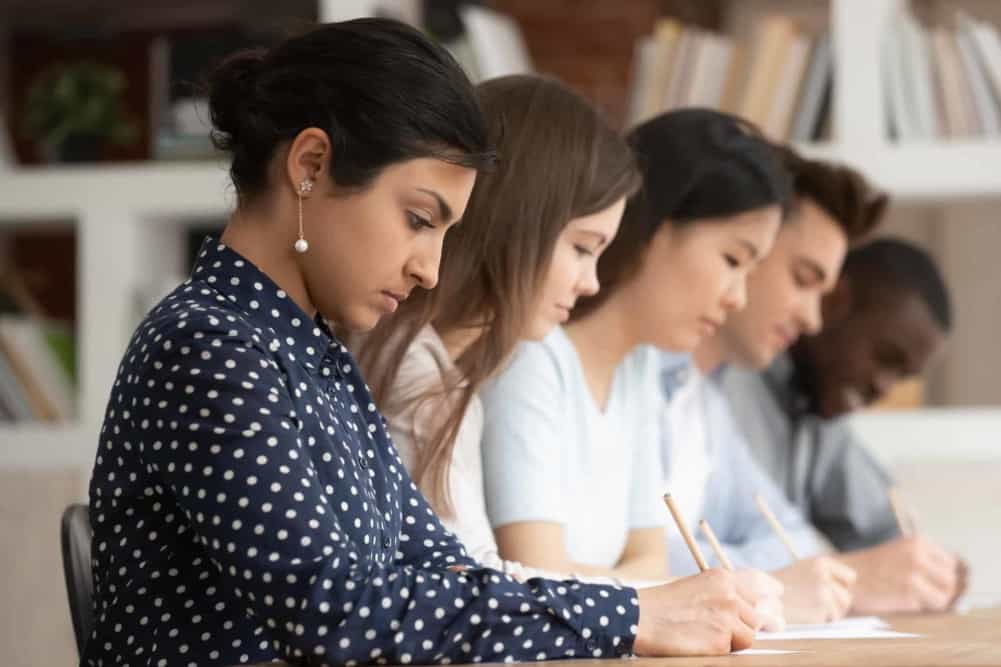 Multi-ethnic concentrated students sitting at desk holding pencils writing on papers doing task, diminishing perspective focus on indian girl study process or assessment - knowledge evaluation concept