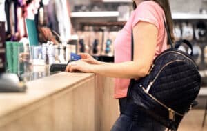 Woman at checkout in fashion store paying with credit card. Customer using payment terminal machine. Standing at counter. Buying and shopping for clothes. Bank card graphics are made up.