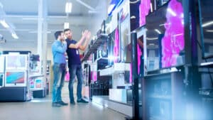 In the Electronics Store Professional Consultant Shows Latest 4K UHD TV's to a Young Man, They Talk about Specifications and What Model is Best for Young Man's Home. Store is Bright, Modern and Has all the Latest Models.