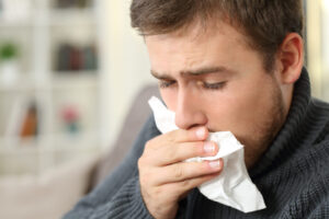 Man coughing covering mouth with a tissue sitting on a couch in a house interior